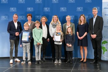 Vier Schülerinnen und Schüler und fünf Lehrerinnen posieren mit Herrn Staatsminister Glauber mit ihren Urkunden.